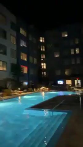 The video depicts an outdoor swimming pool area at night. The pool is illuminated, and the water is still, reflecting the surrounding lights. A multi-story building surrounds the pool, with lights visible in some of the windows. There is a covered structure near the pool with a TV screen visible underneath. The sky is dark and clear. The camera pans across the scene, showing the pool, the building, and the sky. There are no people visible in the shot.