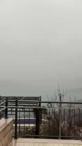 The video shows a view from a balcony overlooking a foggy landscape. The foreground features a metal pergola and railing. A glimpse of a building with a red-tiled roof is visible to the left. The background is obscured by thick fog, with distant shapes of land or islands barely discernible. The atmosphere is overcast and gloomy.