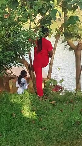 The video takes place outdoors, on a grassy slope adjacent to a body of water, possibly a lake or river. The scene includes trees and other vegetation. Initially, a person wearing red clothing is climbing a tree, with another person in a white and patterned outfit sitting or crouching nearby. A third individual, dressed in dark clothing, is standing near the water's edge, holding what appears to be a mobile phone. The person in red then steps down from the tree and walks towards the individual with the phone.