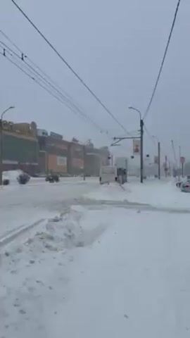 The video shows a snow-covered urban scene on a gray winter day. The foreground features a sidewalk covered in snow, with a large pile of snow separating it from the road. The road itself is also covered in snow, with tire tracks visible. In the background, there is a large, multi-story shopping mall called "GORODOK" in Russian. Several cars are driving on the snow-covered road, moving from left to right in the frame. Streetlights and overhead power lines are visible above the road. Snow is falling lightly, contributing to the overcast and wintery atmosphere. The overall scene depicts a typical snowy day in a city.