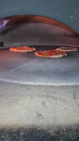 Four small pizzas are arranged on the stone floor of a wood-fired oven. The pizzas are in various stages of cooking, with some showing slightly browned crusts and melted cheese. Flames are visible in the background of the oven, indicating high heat. The camera is positioned close to the oven floor, looking into the oven.