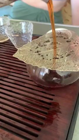 A person is pouring amber liquid from a teapot into a glass cup with a leaf-shaped strainer over it. Two small clear glasses are visible on a wooden tray next to the cup. The tray has horizontal wooden slats and appears to be wet, with liquid pooled in some of the slats. The person's hands are holding the teapot. The background is blurred, but a green garment is visible.