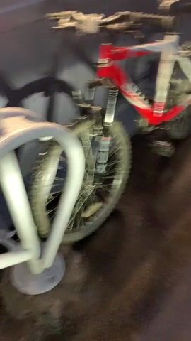 A red and white mountain bicycle is securely parked in a white, U-shaped metal bike rack. The bicycle features a red and white frame with black handlebars, a black seat, and black pedals. The front suspension fork is black with "XCR" visible on it, and the tires have a robust tread pattern. Text including "STS" and the faint outline of "NAVIGATOR" can be seen on the bike's frame.

The bicycle is positioned next to a dark, possibly metallic or painted concrete wall, which casts a clear shadow of the bike's wheels and frame. Above this wall, a brightly lit display window (resembling a vending machine or convenience store window) showcases various products, reflecting light onto the scene.

The ground is dark, paved, and appears wet and highly reflective, suggesting recent rainfall. The overall lighting is dim, indicating either evening or nighttime. There are no people visible in the scene. The setting appears to be an outdoor area adjacent to a building, possibly in a commercial or public space within Касимово-1 Снт, Russia.
