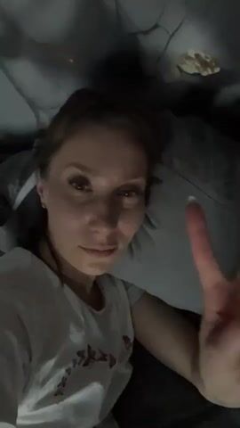 A woman with light brown hair pulled back is lying down and looking at the camera. She is wearing a white t-shirt. Her right hand is raised, and she is making a "peace" sign with her index and middle fingers. In the background, the environment appears to be a bedroom with a pillow and a dark ceiling. The lighting is dim.