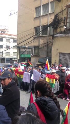 A group of people are gathered in a street, holding Bolivian flags and signs. They appear to be protesting or demonstrating, as they are chanting. The signs have Spanish writing. The street is lined with buildings, and there are numerous overhead power lines. Some of the people are wearing hats, and one person has a "Prensa" shirt. A camera is recording the event. The weather seems sunny.