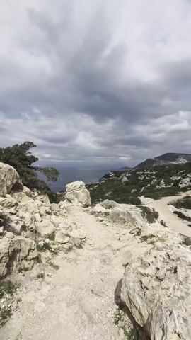 The video shows a rocky, somewhat barren landscape. The foreground is dominated by light-colored rocks and a path, leading towards a view of the sea. In the distance, there's a body of water under a cloudy sky.  A few green trees are scattered throughout the scene, particularly to the left.  On the right, a road winds through a green, hilly area. The overall impression is of a natural, coastal environment under a potentially overcast sky.