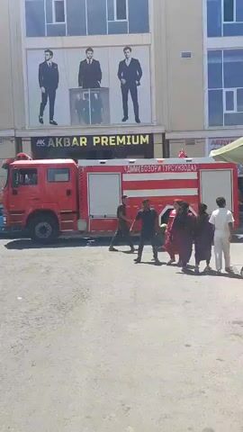 A red fire truck is parked in front of a building with a large advertisement for "AKBAR PREMIUM" featuring three men in suits. Several people, including men and women in traditional clothing, are walking in front of the fire truck and the building. The ground appears to be asphalt. One person on the right is near a small amusement ride.