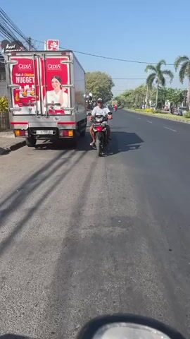 A white box truck with red branding for "KFC" and "Odeya Nugget" is parked on the side of a paved road. The truck has a large advertisement on its side featuring a woman and product imagery. A person is riding a red motorcycle on the road, moving away from the parked truck. The motorcycle has two occupants. The environment appears to be a sunny day with palm trees visible along the side of the road. The road has visible lane markings. The image is taken from the perspective of another vehicle.