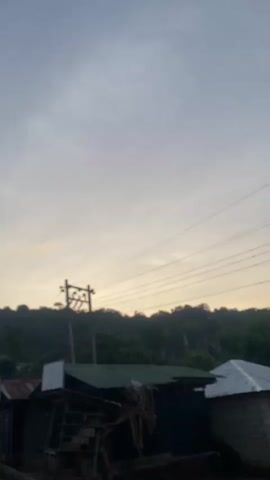 The video presents a static outdoor shot of a rural or semi-urban landscape. The initial frames are out of focus, gradually sharpening over two seconds. A wooden utility pole with multiple overhead power lines stands prominently in the midground. Below the power lines, several simple structures with corrugated metal roofs are visible, some displaying hanging banners or signs, including one with the partially legible text "GAZA." A dense line of dark green trees forms a hillside in the background. The sky is a light grey with a faint yellow-orange hue near the horizon, suggesting either dawn or dusk.