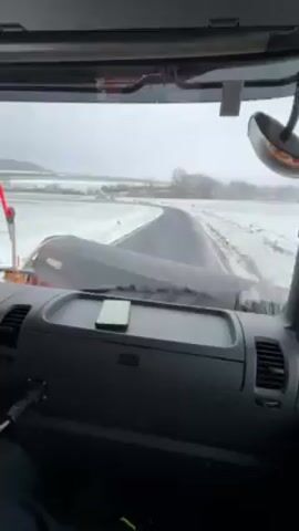 A driver’s point-of-view shows a snow plow vehicle driving down a road. The road is wet and clear, with snow piled on either side. The landscape is flat and snow-covered, with few trees visible. The dashboard of the vehicle is visible.  A phone sits on the dashboard. The wipers of the vehicle are actively moving to clear the windshield of moisture. A red-tipped rod is visible on the left side of the hood. There is some music playing in the vehicle. A sign is visible on the right shoulder of the road. The scene is overcast.