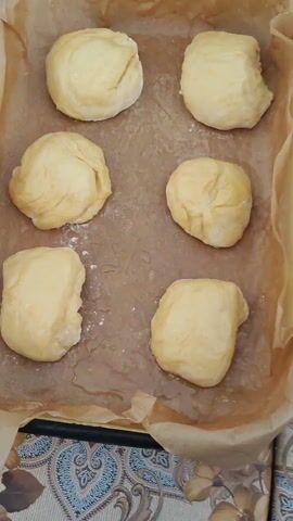 Six uncooked dough balls, yellow in color, are positioned on a baking sheet lined with brown parchment paper. Small pools of water, along with scattered flour, are also present on the paper. The baking sheet sits on a surface featuring a floral pattern. In the background, indistinct audio plays, likely music or speech.