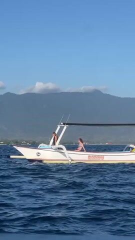 A white motorboat with two people onboard sails across deep blue water under a clear blue sky. The boat has a white canopy supported by a metal frame. A person stands near the front of the boat, and another person sits towards the back. In the background, a green mountainous landscape is visible. The boat creates a spray of water as it moves. The video appears to be shot from another moving boat, as a blurry foreground element suggests a railing or edge.