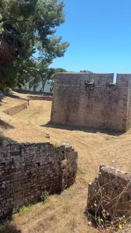 The camera slowly pans across an outdoor scene dominated by crumbling stone walls, possibly ruins or an old fort. The ground is covered in dry grass and weeds, with some greenery growing on and around the walls. The sky is a clear, bright blue. In the distance, a glimpse of blue water can be seen. The architectural style suggests an old, possibly medieval, structure. The overall impression is of a historic site in a state of disrepair.