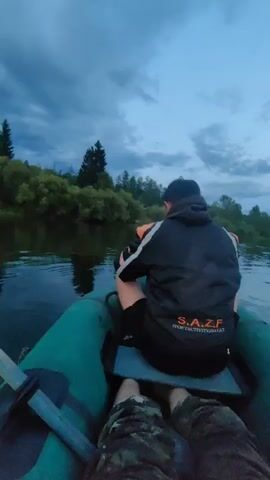 The video captures a point-of-view shot from a boat on a river at dusk. In the foreground, the green inflatable side of the boat is visible, along with a portion of a person's legs wearing camouflage pants. In the middle ground, a person wearing a black hooded jacket with white accents and black shorts is seated in the boat, facing away from the camera. The jacket has a logo on the back that reads "S.A.Z.F. SPORTACTIVITY.COM". An oar with a wooden handle and a dark paddle is also visible in the boat. The river is calm, reflecting the cloudy sky. The background features a lush green treeline along the riverbanks, with darker green trees dominating the left side and lighter green foliage on the right. The sky is filled with dramatic, dark clouds, with a hint of orange and yellow light peeking through on the horizon, suggesting either sunrise or sunset. The overall atmosphere is serene and natural.