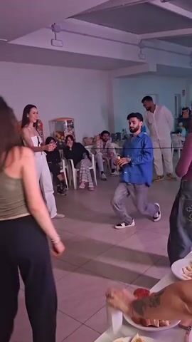 A group of people are gathered indoors in a casual setting. Several individuals are seated in chairs, and others are standing around watching a man in a blue shirt and gray pants do the limbo with a bottle in his hand. The room has a tiled floor and white walls. There is a table with food in the foreground, and an individual in a white outfit standing in the background. The lighting appears to be a mix of natural light and artificial light, possibly from overhead fluorescent fixtures. The overall atmosphere seems relaxed and festive, suggesting a social gathering or party.
