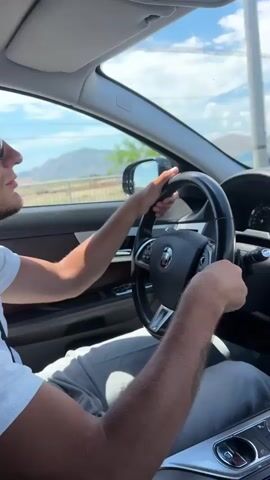 A person is driving a car. The camera is inside the car, looking forward. The driver's hands are on the steering wheel. The dashboard of the car is visible, including the navigation screen. Outside the car, mountains and trees are visible under a cloudy sky. The car appears to be moving on a road.