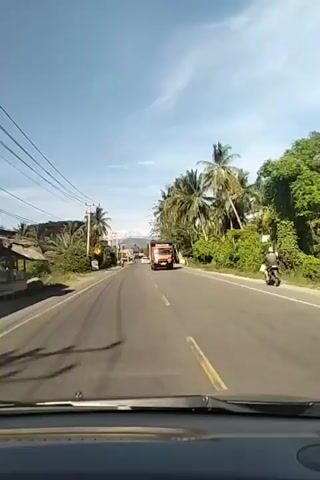 The video shows a view from inside a vehicle traveling down a road lined with palm trees and lush greenery under a clear blue sky. The road has a yellow dividing line and appears to be paved. Several vehicles, including a large orange truck, two white cars, and some motorcycles, are visible on the road ahead. Power lines run along the left side of the road, supported by utility poles. In the distance, mountains can be seen. The camera perspective suggests the video is a first-person recording from a driver's point of view.