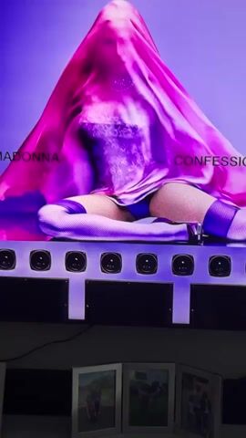 The video shows a person draped in a pink and purple veil, sitting on a surface with multiple speakers. The person is wearing thigh-high stockings and appears to be partially clothed underneath the veil. Text is visible on the veil, reading "MADONNA CONFESSION." The lighting is predominantly purple and pink.