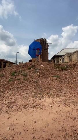 A young person stands on a mound of dirt in what appears to be a rural village. The person is covered in gray paint and is wearing a red wig and a red skirt. They are holding a bright blue umbrella over their head. Behind them is a dilapidated mud brick structure. Other simple buildings can be seen in the background under a partly cloudy sky. The person is stationary as the camera approaches.