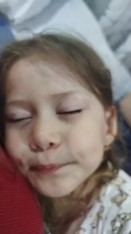 The video is a close-up shot of a young girl who appears to be sleeping or resting with her eyes closed. She is lying down, possibly in bed, and her head is resting on someone's shoulder, indicated by the red fabric visible on the left side of the frame. The girl has light brown hair and is wearing a white shirt with a pattern of cartoon bears. She has a small gold earring in her right ear. Her facial expression is relaxed. The lighting suggests it might be daytime or an indoor setting with artificial light. The overall mood is peaceful and intimate. The video is in Russian.