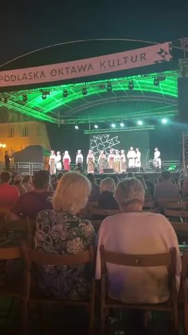 A video displays a live outdoor performance with a stage illuminated by green lights. A banner across the top reads "PODLASKA OKTAWA KULTUR". On stage, a group of performers, mostly women dressed in traditional folk costumes, stand in a line. They appear to be singing or performing. In the foreground, the backs of two audience members are visible, seated in chairs facing the stage. The overall environment suggests a cultural festival or event held at night.