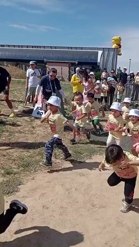 The video shows a group of children and adults in an outdoor setting with a dusty, dirt ground and sparse vegetation. Several children, appearing to be young, are running and playing. One adult is holding a camera on a gimbal, filming the scene. Other adults are standing and watching. In the background, there are some structures and more people. The overall atmosphere seems to be one of a casual outdoor event or gathering, possibly a race or a fun activity for children. The lighting suggests it is daytime with a clear sky.