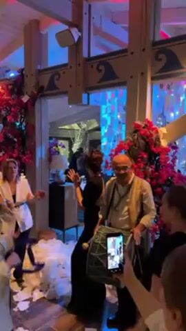 The video shows people at an indoor celebration. A man with a shaved head plays a large drum decorated with green material. A woman in a black dress claps her hands. The scene has flowers, decorations, and colored lighting. Some people hold up their phones to record the scene. Some objects can be seen through a glass door in the background.