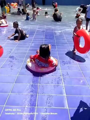 A group of children and adults enjoy a sunny day at a water park. The pool has a tiled floor and is filled with people, many using inflatable rings for support. Several children are visible on the tiled deck near the water, handling more inflatable rings. One boy lies back on an inflatable while a younger girl stands beside him, looking at him with curiosity. The atmosphere appears lively and playful.