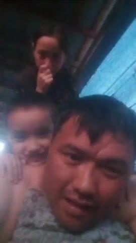 The media file is a selfie video of a man with two children on his shoulders. The man is in the foreground, looking at the camera with a slight smile. He is wearing a patterned shirt. The two children are on his shoulders, with the younger child in front and the older child behind. Both children are smiling and looking towards the camera. The younger child has his arms around the man's neck, and the older child has her arms around the younger child's shoulders. The background is dimly lit and appears to be an indoor setting, possibly a home, with wooden beams visible overhead. The lighting suggests it could be daytime, but the indoor setting makes it difficult to determine the exact time of day. The overall mood of the video is happy and familial, with the subjects appearing to be enjoying themselves. The location context provided is Іргелі ауылдық округі, Kazakhstan, suggesting this is where the video was taken. There is no visible text in the media file.