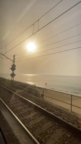 This is a short video clip of a train journey, taken from the perspective of a passenger looking out the window. The sun is shining brightly, creating a glare on the water, and illuminating the sky with a hazy, golden light. The sea is calm, with gentle waves lapping at the shore. In the foreground, there are train tracks, and a fence separates the tracks from the beach. A few people can be seen walking on the beach, and there are also some trees and vegetation along the coastline. The video is taken during the daytime, likely in the morning or late afternoon, given the angle of the sun.