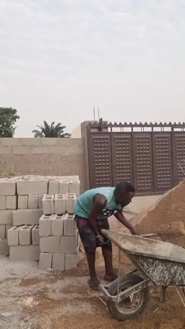 A man is working on a construction site. He's wearing a teal tank top, patterned shorts, and sandals. He uses a shovel to fill a wheelbarrow with sand from a large pile. Behind him is a stack of grey concrete blocks and a tall brown metal gate. The sky is overcast.