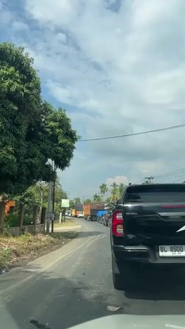 The video shows a view from inside a vehicle driving on a two-lane road with traffic. A black pickup truck is in the foreground. The road is lined with trees and vegetation on the left side. In the distance, there are more vehicles, including several trucks, creating a traffic jam. The sky is partly cloudy.