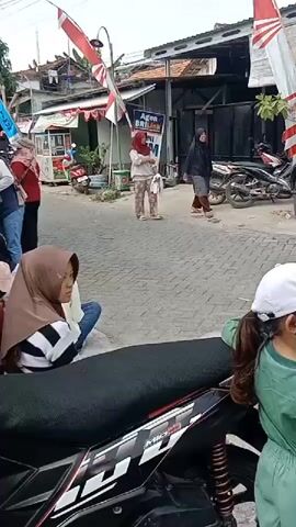 A medium shot captures an outdoor scene on a paved street. In the foreground, a young girl wearing a white cap and a green tracksuit is leaning against a black and white motorcycle. She is looking to the right. To her left, a woman wearing a brown hijab and a striped shirt sits on the ground, also looking to the right. Behind them, the street continues with several people standing and walking. Two women are in the middle ground: one in a light-colored hijab and patterned pants, and another in a black hijab and grey dress. They appear to be conversing. Several motorcycles are parked along the street, and buildings with red and white Indonesian flags are visible in the background. An ATM sign for BRI Link is also present. The overall atmosphere appears casual and everyday.