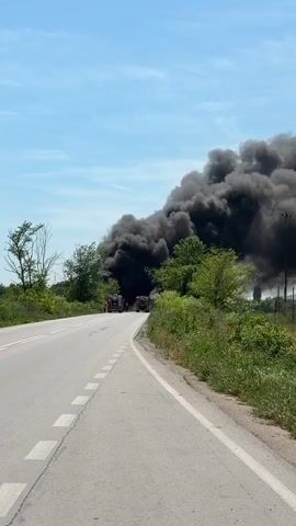 The video shows a dark, thick plume of smoke rising into the sky from an unspecified source. A road is visible in the foreground, and a truck is parked on the side of the road. Trees and other vegetation are also visible.