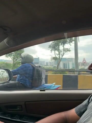 The video shows a view from inside a vehicle, looking out the side window. The perspective is dynamic, suggesting the vehicle is in motion. In the foreground, a black and yellow striped barrier is visible, indicating a road or highway. Beyond the barrier, several modern buildings can be seen in the distance, some appearing to be office complexes or high-rises. Trees are scattered throughout the landscape. In the middle ground, a motorcycle rider wearing a blue shirt and a grey helmet is visible, carrying a black backpack. Further ahead, a white car with a yellow license plate (TG 26 T 4792) is also in motion. The sky is overcast with clouds. The interior of the vehicle is partially visible, including a side mirror reflecting the road behind, and parts of the dashboard and car door.