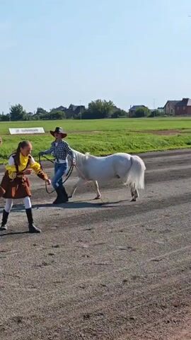 The video shows a white pony being led by a person in a cowboy hat. Two individuals dressed in brown skirts and yellow tops are crouched behind the pony. They are all on a dirt or gravel surface, possibly a track. In the background, there is a green field with trees and houses. The sky is blue and clear. The person in the cowboy hat is holding a lead rope. The two individuals behind the pony appear to be interacting with or adjusting something on the pony.