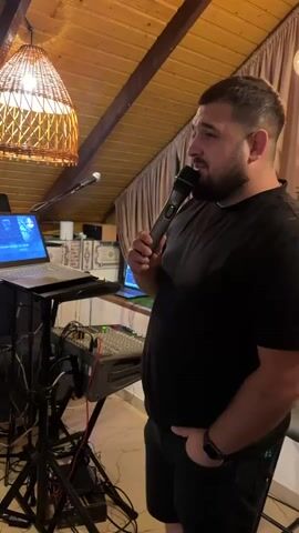 A man wearing a black t-shirt and black shorts holds a microphone and sings. He is standing in front of a laptop and a mixing board on a stand. The scene is indoors, with a sloped wooden ceiling and a woven lamp hanging from it. There are curtains in the background.