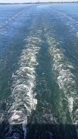 The video shows a view from the back of a boat moving through the water, leaving behind two distinct wakes. The water is a deep blue-green and choppy due to the boat's movement, with white foam and turbulence creating textured patterns. In the distance, a hazy shoreline is visible under a clear blue sky, suggesting a sunny day. The shoreline features what appear to be industrial structures or buildings, hinting at an urban or port area. The overall scene conveys a sense of travel and the vastness of the sea. There is no visible text in the media file. The location context provided is Helsinki, Finland.