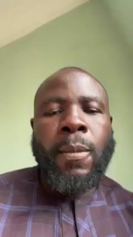 A man with dark skin, a full beard, and a maroon and blue plaid shirt is centered in the frame against a light green wall. He holds a toothpick with a small, dark piece of food on it, and he puts the food into his mouth and chews.