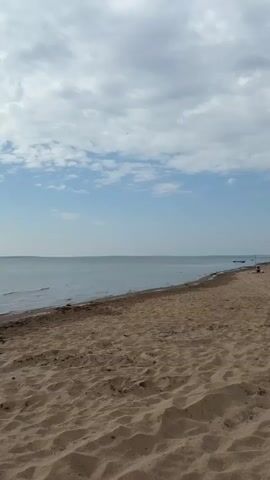 A video displays a sandy beach with the ocean in the background and a cloudy sky above. The camera is positioned low to the ground, looking out towards the water. The sand is light brown and has ripples from wind or water. The ocean is calm and appears to be a light blue-grey color. The sky is a bright blue with scattered white clouds. There are no people or objects clearly visible in the foreground or midground. In the far distance, a thin line of land can be seen along the horizon, suggesting a distant coastline.