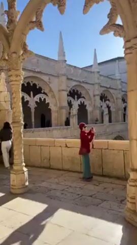 The video shows a woman in a red sweater and green skirt standing on a stone walkway, possibly a balcony or terrace, and taking a photo with her phone. She is facing toward the center of a courtyard or cloister, framed by ornate stone arches and columns. In the background, there is a large, light-colored stone building with multiple archways and spires. Several other people are visible in the background, walking along the upper levels of the building. The sky is clear and blue. The light suggests it is a sunny day, casting shadows from the arches and columns onto the walkway. A second person appears on the right edge of the frame, dressed in dark clothing.