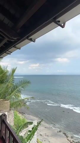 The video pans over a tranquil ocean scene with a cloudy sky. In the distance, a pink lighthouse stands in the water. Palm trees line the shore in the foreground. The camera is positioned on a balcony or raised platform, looking down and out towards the sea. The waves gently crash on the dark sand beach.