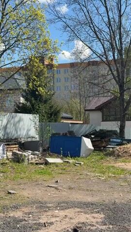 The camera pans across a messy outdoor yard in daylight. The ground is a mixture of dirt, gravel, and sparse grass. In the foreground are piles of construction materials like bricks and concrete slabs, along with a blue and white container lying on its side. Behind these items is a corrugated metal fence. Beyond the fence, a small building with a red tile roof is visible, partially obscured by trees. Further back is a large, light-colored apartment building. The most prominent feature of the yard is the presence of two disassembled trucks, their cabs open and parts scattered around a mound of sand. The sky is blue with scattered clouds, and the surrounding trees are just starting to leaf out. The scene conveys a sense of disrepair and ongoing construction.