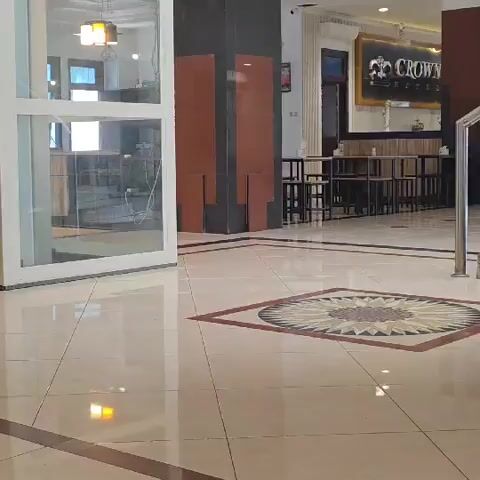 This is a low-angle, wide shot of an indoor lobby area. In the foreground, the polished tile floor has a geometric inset pattern. To the left, there is a large, glass-enclosed elevator, next to a dark wood display cabinet with glass doors, filled with trophies. A pillar with brown and red paneling is visible to the right of the elevator, and beyond that, a seating area with tables and chairs. A sign with a crown emblem is mounted on the wall above the seating area. The lighting is dim, with reflections visible on the floor.