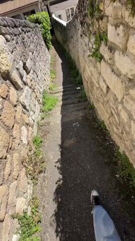 The video opens with a downward view of a narrow stone pathway or staircase, flanked by old stone walls with some greenery growing on them. Houses with dark tiled roofs are visible at the bottom of the path. The camera then pans upwards to a bright, clear blue sky with a strong sun glare. Following this, the camera pans left to reveal a historic-looking stone church or cathedral with a prominent steeple, situated on a hill or elevated ground. Other houses with tiled roofs are in the foreground, and trees are visible in the distance. The camera then pans back to the right, showing the bright sky and sun again, before returning to the initial view of the stone pathway and houses. The scene is brightly lit under a clear, blue sky on what appears to be a sunny day in Saint-Leu-d'Esserent, France. No people or text are visible.