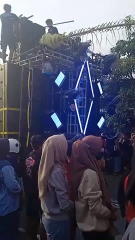 A daytime, slightly low-angle shot captures a lively outdoor event. A crowd of people, mostly young, stand in the foreground, many wearing casual clothing like hoodies and jeans. Several young women in the foreground are wearing hijabs. Behind them stands a large sound system, constructed on a metal frame. The system features a central diamond shape formed by bright blue lights. Above the lights are more speakers and decorations made of bamboo. People are visible on top of the speaker system, adjusting equipment. The background includes trees and glimpses of buildings. The overall atmosphere suggests a festive or celebratory event, with the sound system as a focal point.