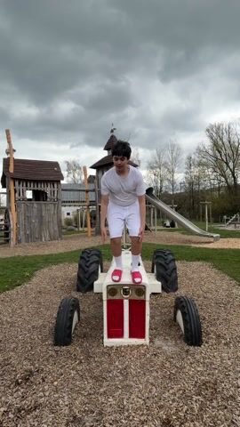 A young male teenager in a white t-shirt, white shorts, and red sandals jumps in the air on top of a small, white, toy tractor with a red grill. The tractor is situated on a bed of brown wood chips in a playground. The teenager is facing forward with arms outstretched. In the background, there are wooden playground structures including a tower and a slide, as well as trees and overcast skies.