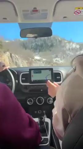 The video shows an interior view from the front seats of a vehicle.  The dashboard is visible, featuring a navigation screen displaying a route. The driver, wearing a dark purple top, has their hands on the steering wheel. The passenger, wearing a light-colored sweatshirt, has long brown hair that blows across her face. The car is driving on a mountain road, and through the windshield, a snow-covered landscape is visible under a clear blue sky.  Music is playing in the background.