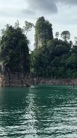 This media file captures a scenic view from a boat on a body of water, likely a lake or reservoir, given the presence of large limestone karsts. The karsts, which are prominent in the foreground and background, are covered in lush green vegetation, with trees growing even from the sheer rock faces. The water is a deep, vibrant turquoise-green, with gentle ripples reflecting the overcast sky.

The time of day appears to be daytime, possibly late morning or afternoon, as indicated by the diffused light and the sky being partly cloudy. The weather seems to be mild, with no harsh sunlight visible.

In the foreground, the back of a person's head, wearing a black baseball cap, is visible, suggesting the viewer is on a boat and observing the landscape. There are also some submerged, dead tree trunks or branches visible in the water, a common sight in reservoirs formed by damming rivers.

The overall atmosphere is serene and majestic, highlighting the natural beauty of the location. The towering karsts and the tranquil water create a sense of awe and wonder. The scene evokes a feeling of exploration and immersion in nature.

While there is no visible text in the media file itself, the location context provided is Khao Phang, Thailand, a region known for its dramatic limestone formations and scenic waters, such as Cheow Lan Lake in Khao Sok National Park. This context helps to identify the specific type of landscape being depicted.