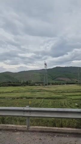 The video shows a landscape view from a moving vehicle, likely a car, looking out over green fields and hills under a cloudy sky. A metal guardrail is visible in the foreground, separating the road from the fields. In the middle ground, a tall metal electricity pylon stands amidst the green landscape. The hills in the background are covered with trees and vegetation, with some evidence of terracing or cleared areas. The overall impression is of a rural or agricultural setting.